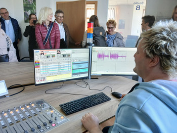 Radiostudio mit Person an Informations- und Tontechnik im Vordergrund und mehre-ren Personen im Hintergrund