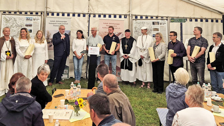 große Gruppe von Personen im Halbkreis vor Publikum stehend, teilweise in mittelalterlichen Kostümen von Geistlichen und Pilgern