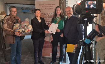 Drei männliche und eine weibliche Person bei der Übergabe eines Bescheides und Präsentkorbes