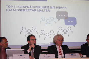 Zwei Männer auf Bühne vor einer Präsentation, einer spricht in ein Mikrofon