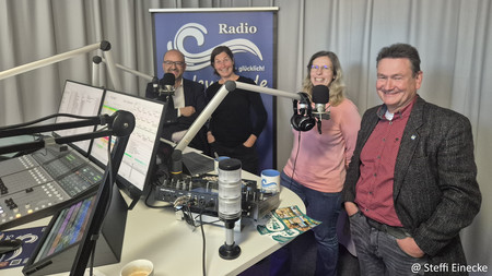 2 Männer und 2 Frauen stehen in einem Radiostudio und lächeln in die Kamera