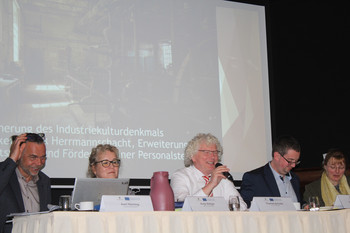 5 Personen an langem Tisch auf Bühne mit Präsentation im Hintergrund